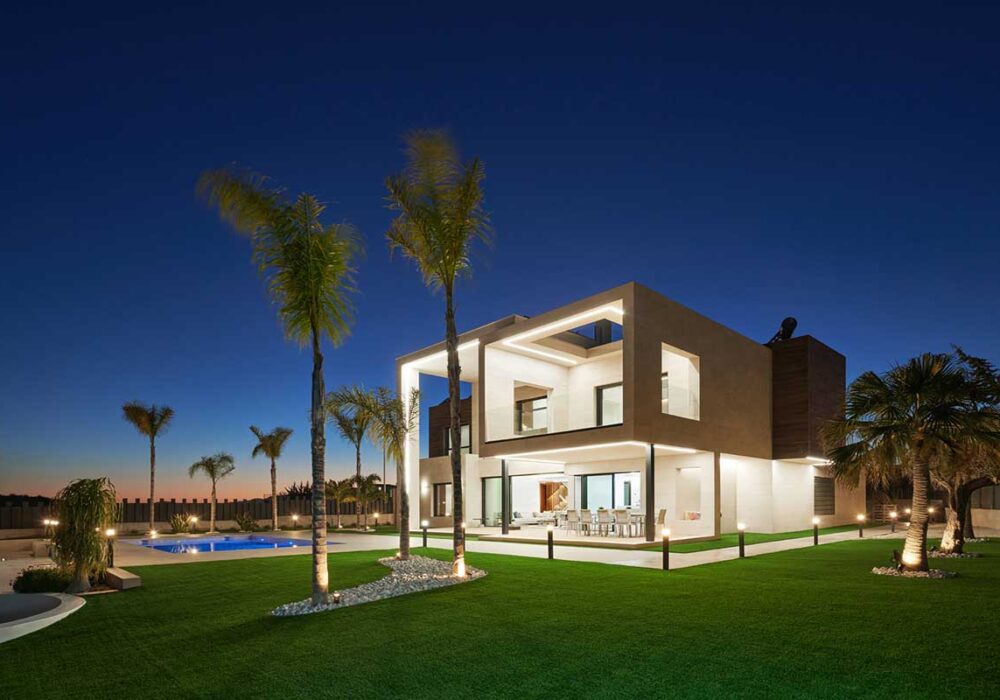 Arquitectos Murcia Proyecto Czech-Rizqy. Vista nocturna de la casa desde el jardín con dos palmeras de frente.